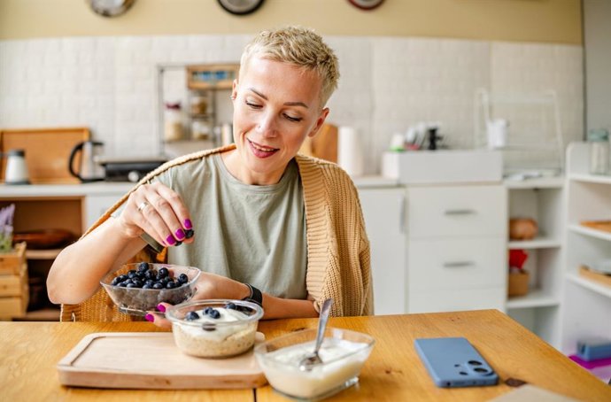 Archivo - Mujer desayunando en la cocina arándanos con yogur.