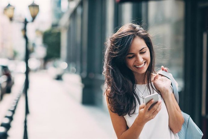 Archivo - Smiling young woman walking outdoors at urban setting and checking messages.