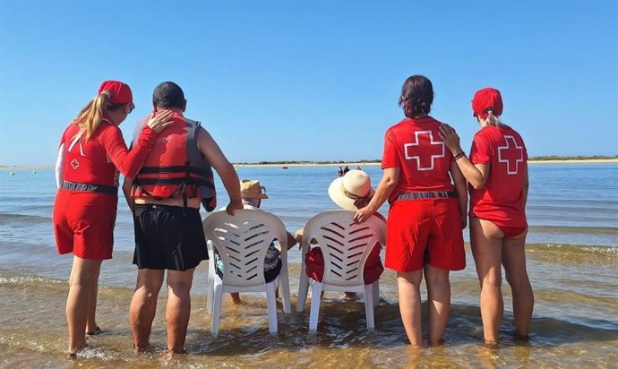 Miembros de Cruz Roja Huelva acompañan a dos personas mayores en la playa.
