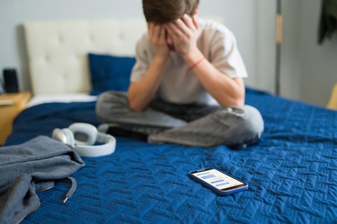 Archivo - Adolescente triste sentado en su cama después de consultar sus redes sociales en el móvil.