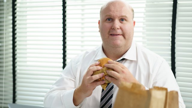 Archivo - Hombre con sobrepeso comiendo hambuerguesa en la oficina, mirando a la cámara.