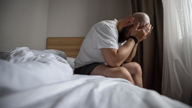 Archivo - Hombre sentado en la cama sujetándose la cabeza en una mañana penumbra en el dormitorio. Estrés, depresión, soledad