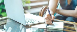 Archivo - Mujer escribiendo a mano con papel y bolígrafo junto a un ordenador portátil.